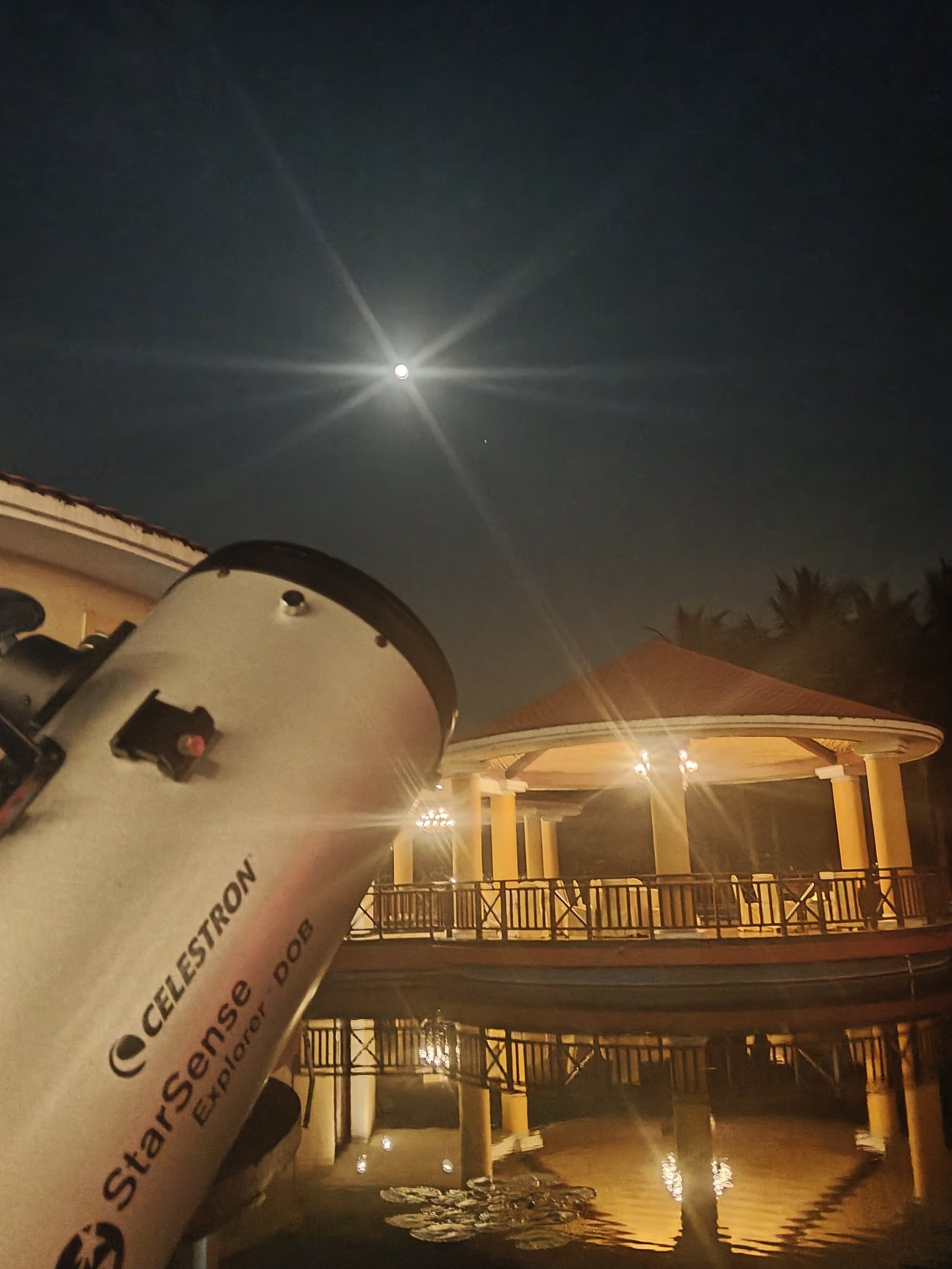 A Celestron telescope stands on the observatory deck at night beneath a brilliant star, with the lit observatory building behind it.